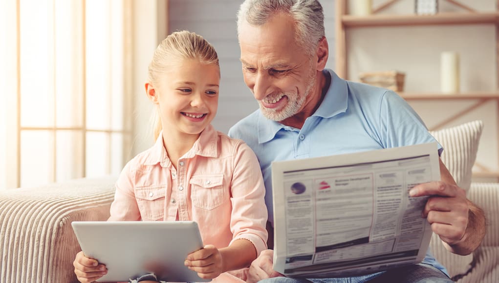 Grandpa and granddaughter on tablets reading news from newspaper and tablet, respectively