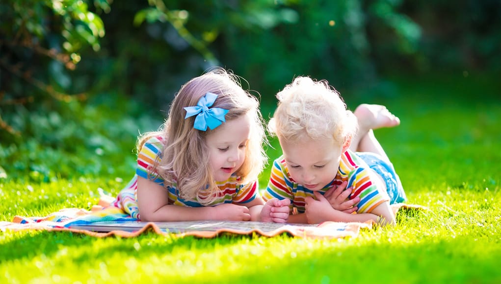 2 young preschool kids engaging in outdoor learning and place-based ed by doing homework and reading outside