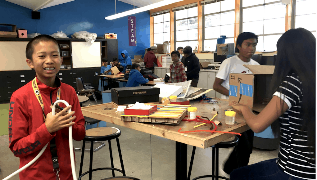 A student standing in a makerspace classroom at VIDA, holding some sort of tube contraption and smiling