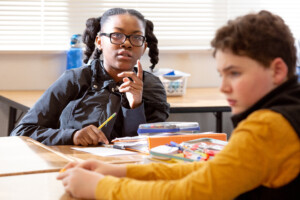 Two middle school students collaborate at a desk, one gesturing thoughtfully while explaining an idea to her classmate.