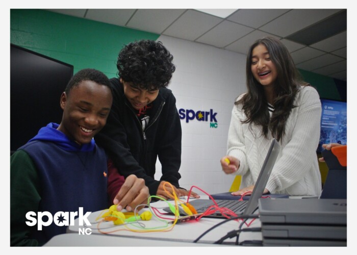 Three diverse students laughing and collaborating on a hands-on tech project at a SparkNC learning space.