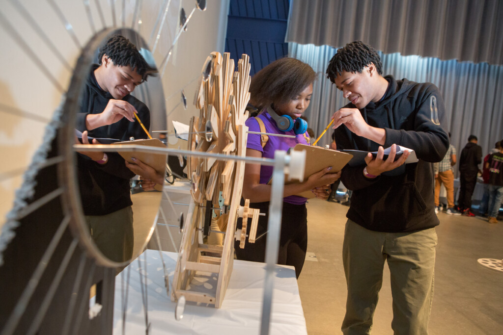 Three students taking notes on clipboards while closely examining a wooden mechanical model at a STEM exhibit.