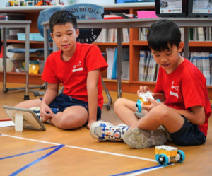 Two students in red school uniforms program a small robot together on a classroom floor using a tablet.